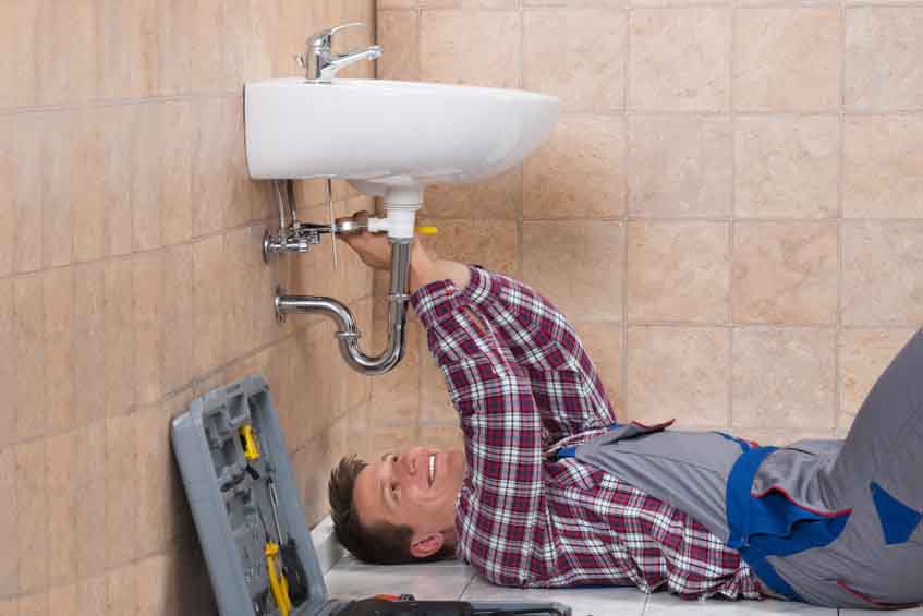 Happy Young Male Plumber Lying On Floor Fixing Sink In Bathroom