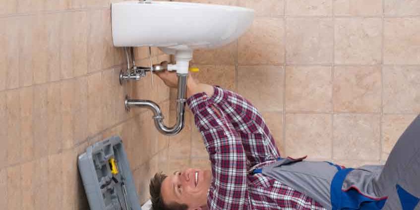 Happy Young Male Plumber Lying On Floor Fixing Sink In Bathroom